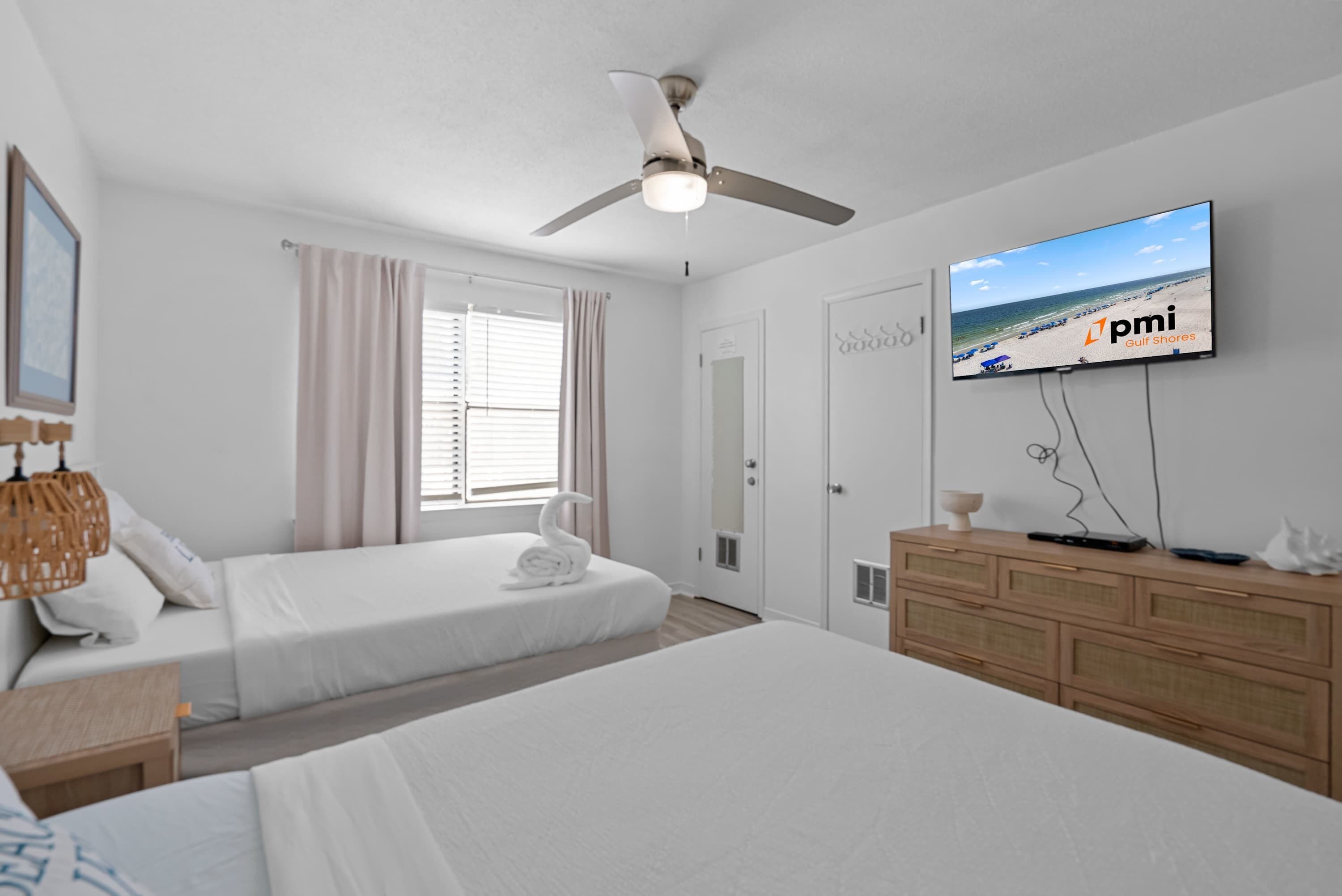 Hotelkamer met twee bedden, een plafondventilator, een televisie waarop een strandscène te zien is, en een dressoir.