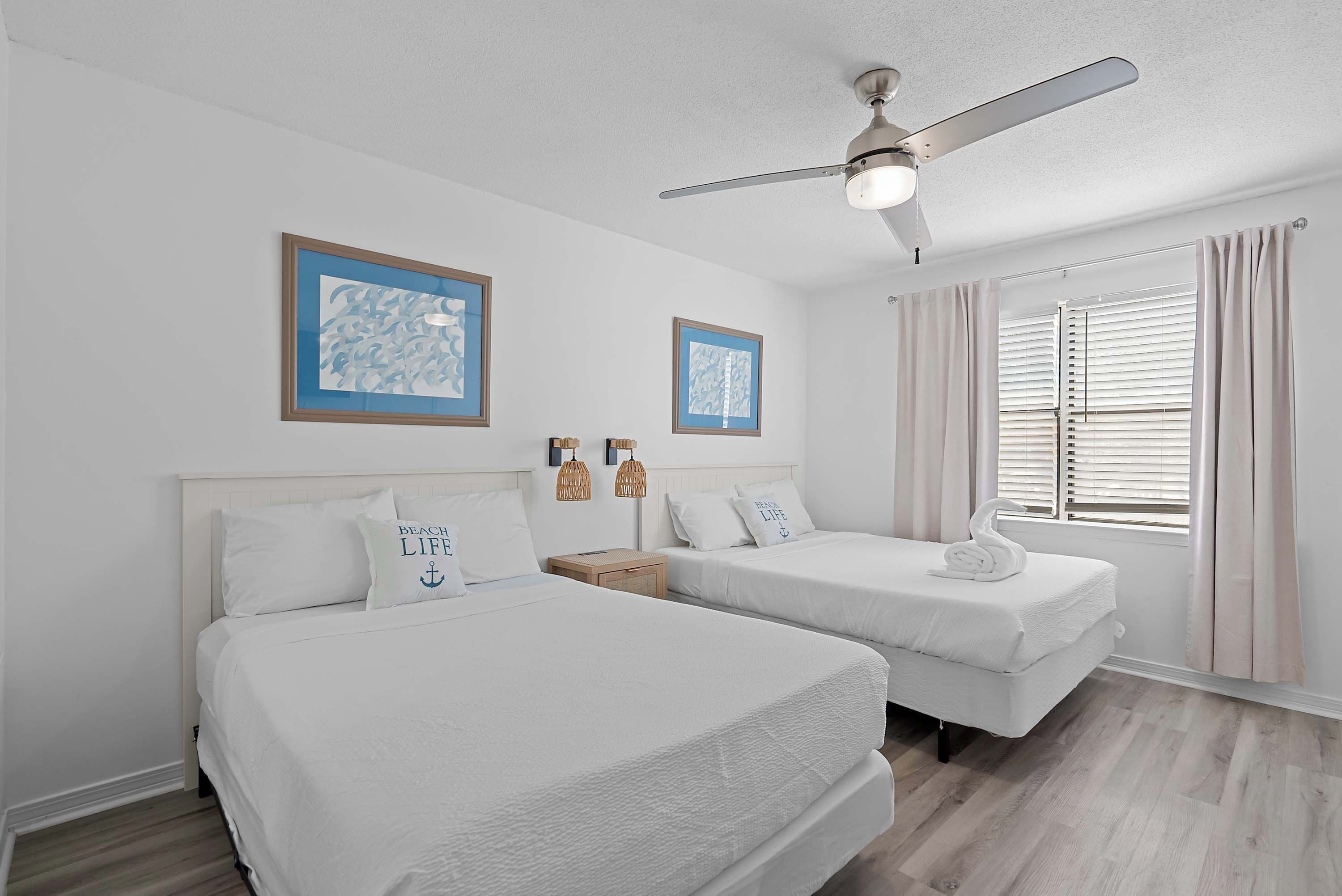 A hotel room with two beds, a ceiling fan, and two framed pictures on the wall.