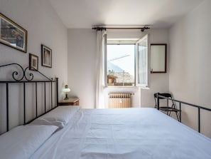 A bedroom with a bed, a nightstand with a lamp, a window with curtains, and a view of buildings outside. - Apartment Ilda by Interhome (Morgex)