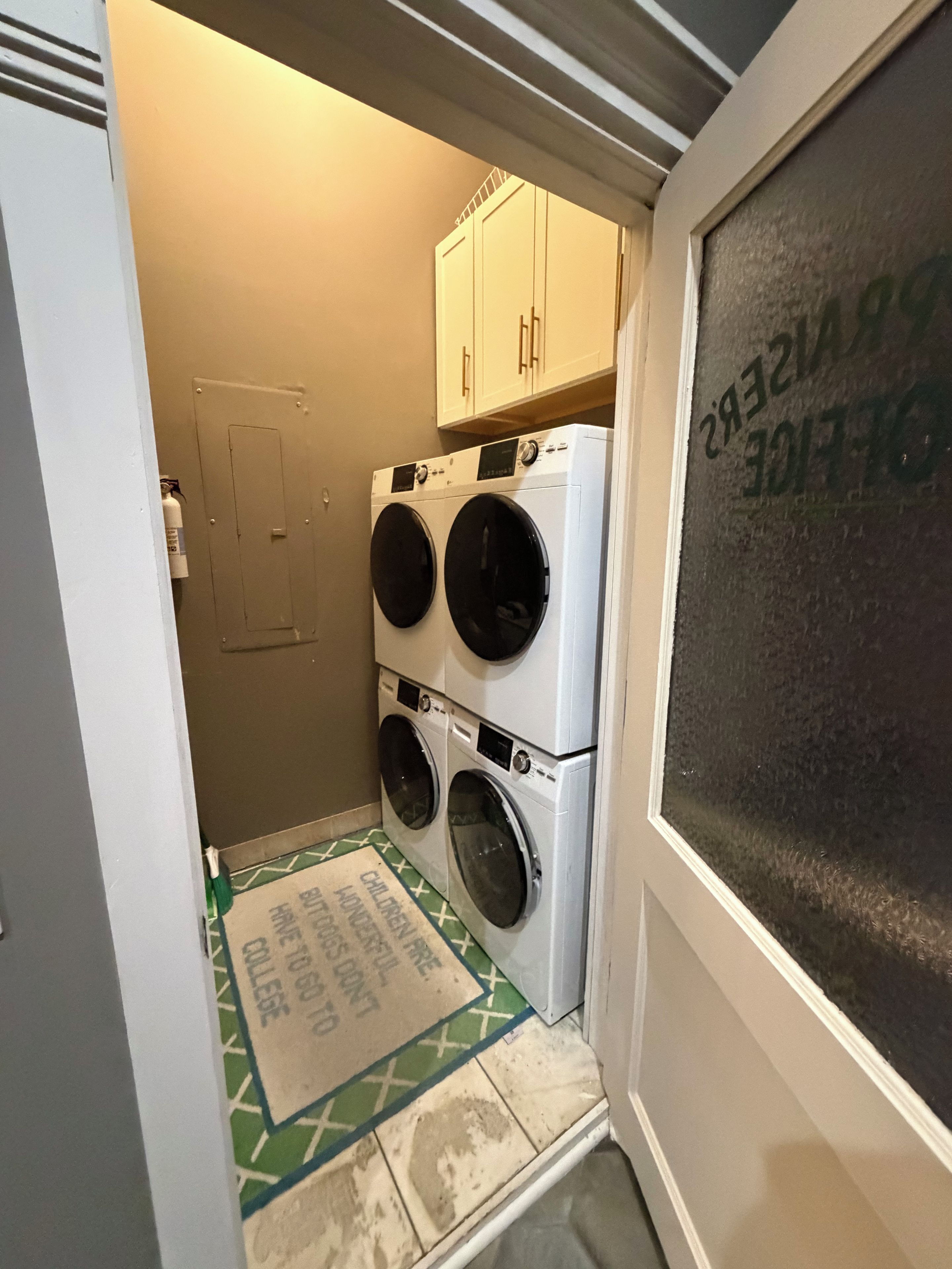 A laundry room with stacked washing machines, a door, and a mat with text.