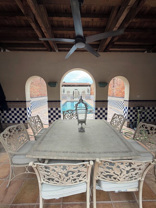 Un patio techado con mesa y sillas, un ventilador de techo y vistas a un edificio con arcos.