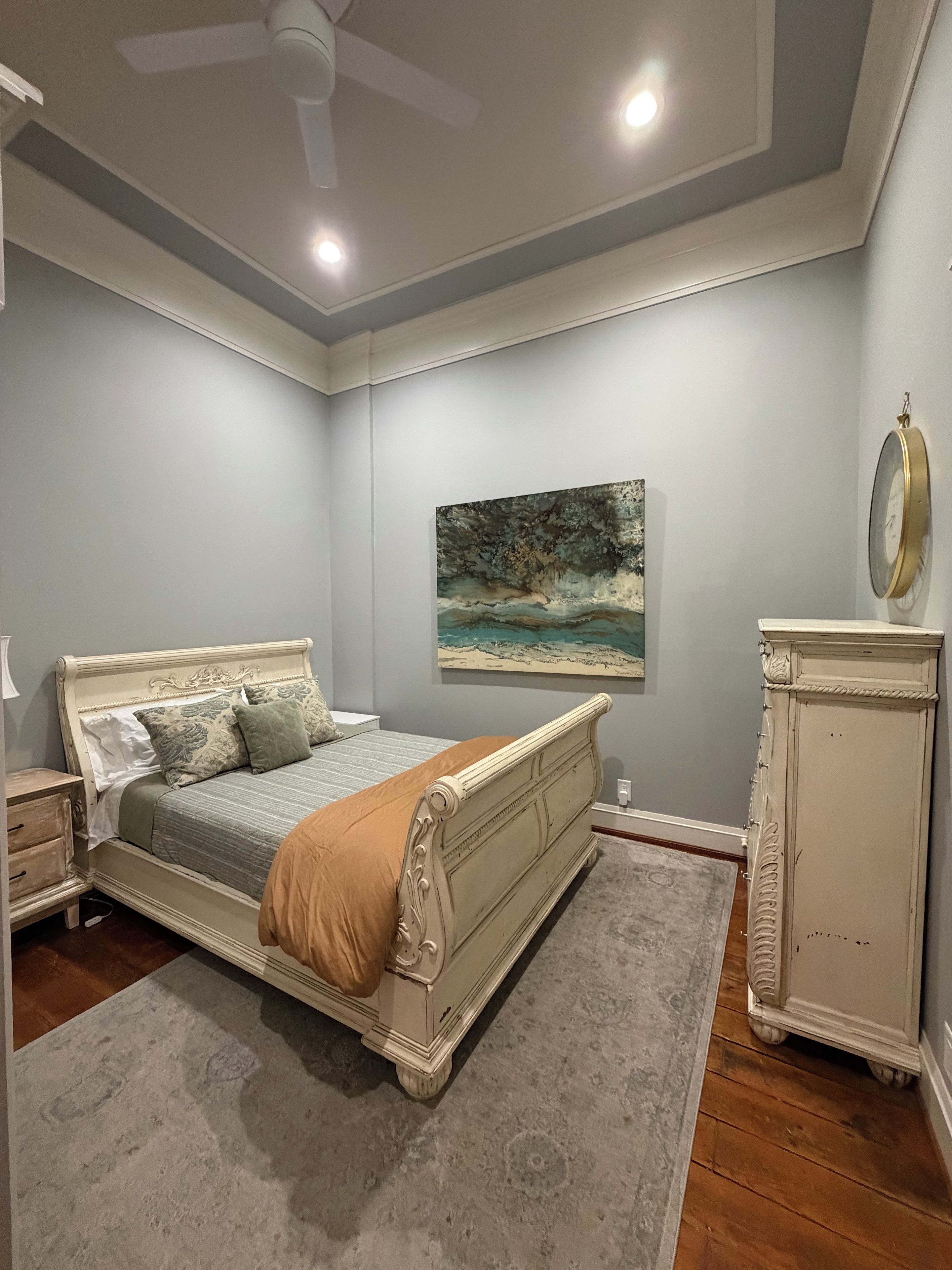 A bedroom with a bed, a nightstand, a dresser, a ceiling fan, and a painting on the wall.