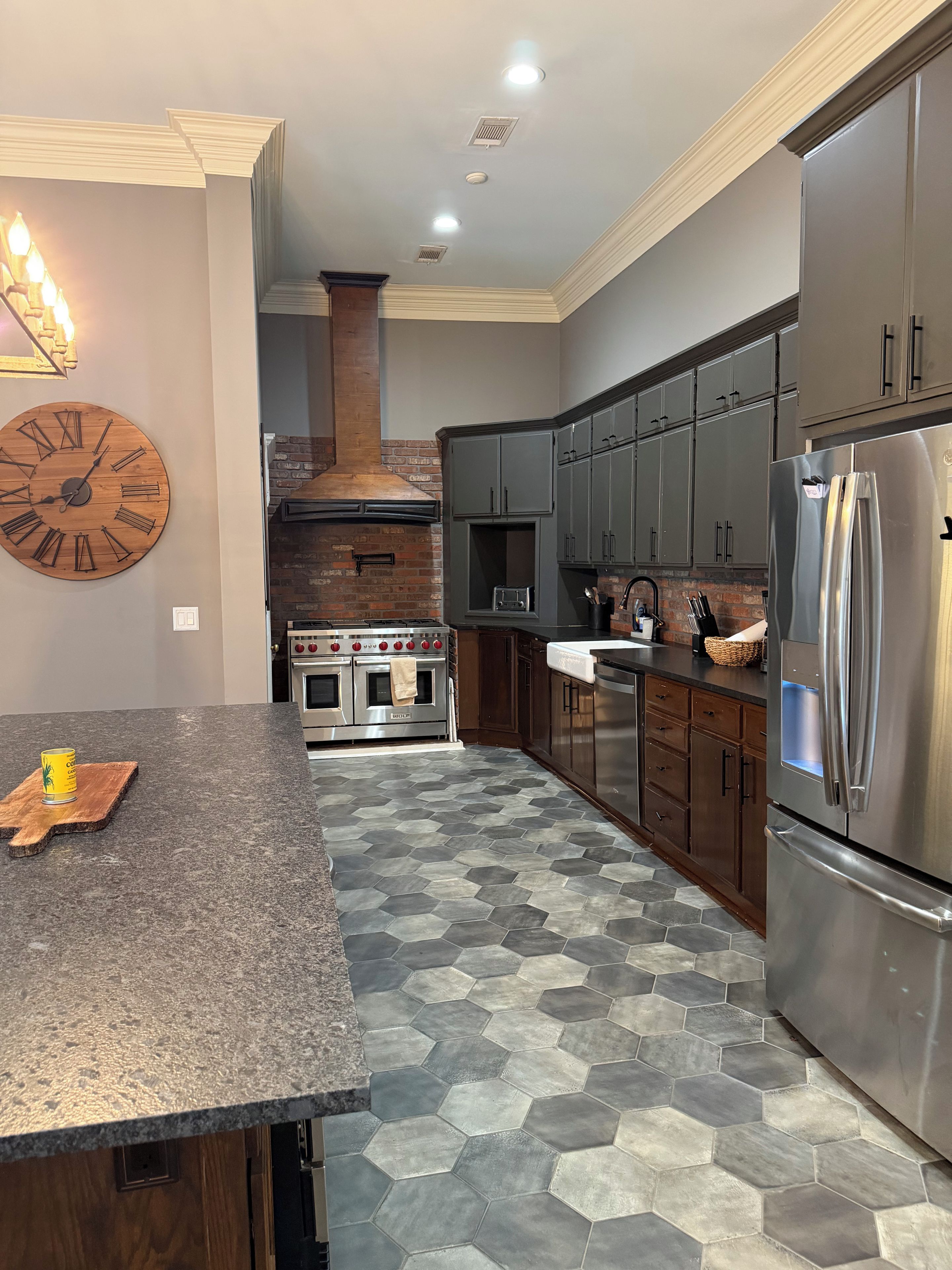 A kitchen with a large wooden clock, stainless steel appliances, and hexagonal tile flooring.