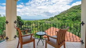 A balcony with two wooden chairs and a small round table, overlooking a lush green landscape and the sea. - 3BR Retreat | Infinity Pool | Sunsets (Las Catalinas)