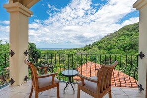 Ein Balkon mit zwei Holzstühlen und einem kleinen runden Tisch, der auf eine üppige grüne Landschaft und das Meer blickt.
