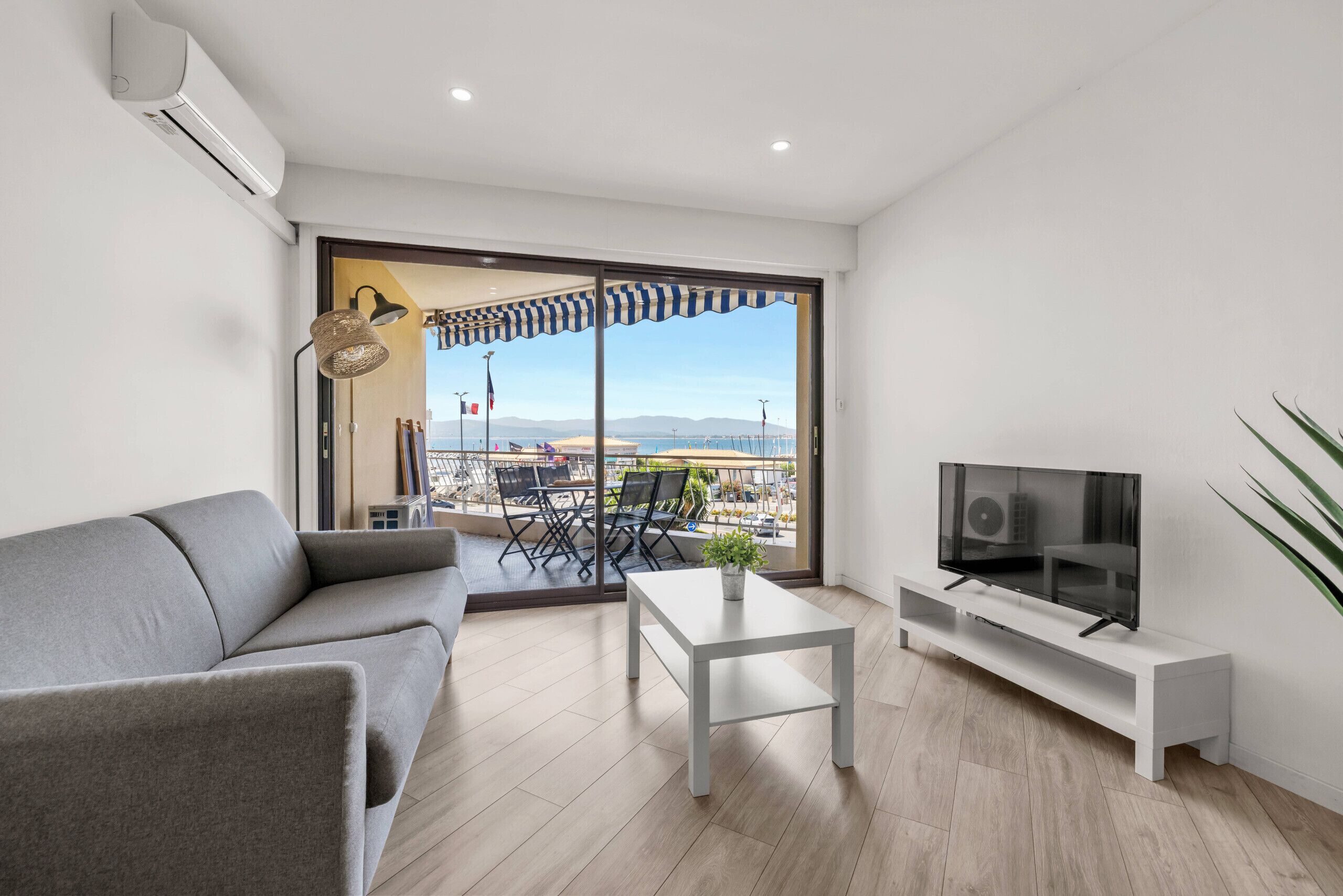 A modern living room with a grey sofa, a coffee table, a flat-screen TV on a white stand, and a large sliding glass door leading to an outdoor area with a view of the sea and mountains.