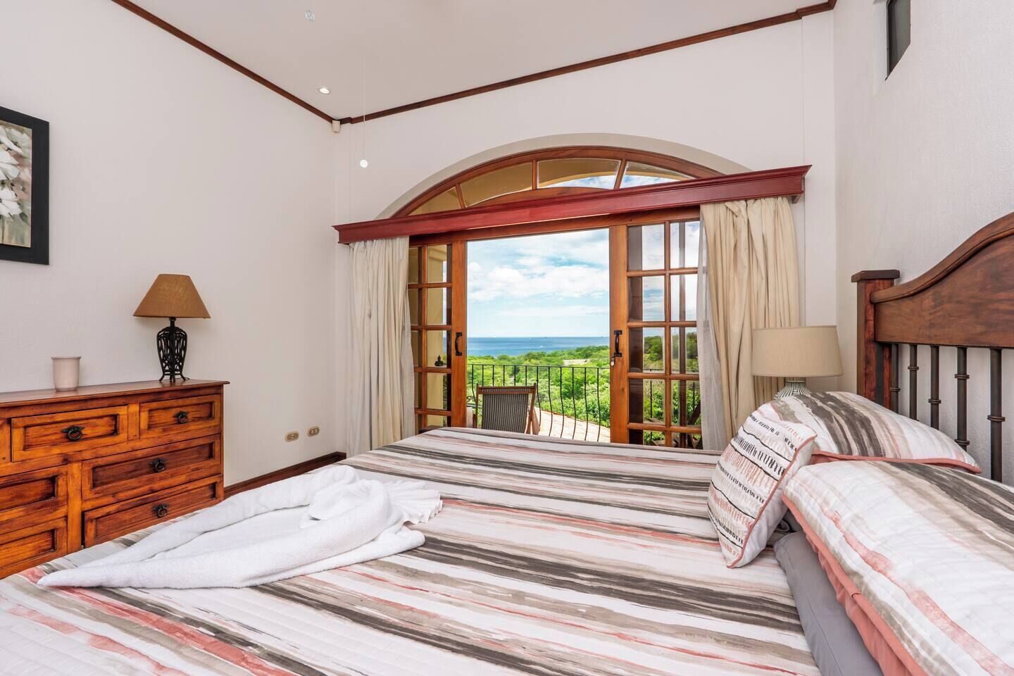 A bedroom with a large bed, wooden dresser, and a balcony with a view of the sea.