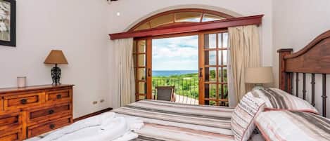 A bedroom with a large bed, wooden dresser, and a balcony with a view of the sea.