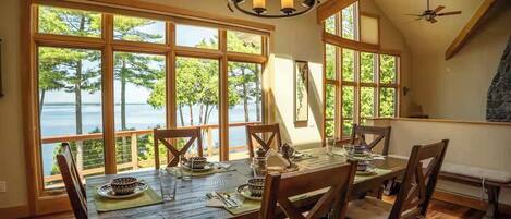 Un comedor con una mesa dispuesta para cuatro, ventanas amplias con vistas a árboles y agua, y una chimenea de piedra.
