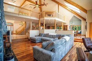 A spacious living area with a large sectional sofa, a wooden staircase, and a high ceiling with exposed beams.