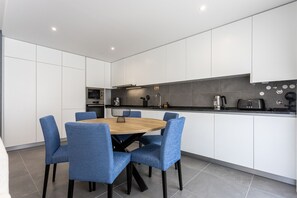 A modern kitchen with white cabinets, a black countertop, and a round wooden table with blue chairs. - Galeão Sea Apartment - BY SCH (São Martinho do Porto)