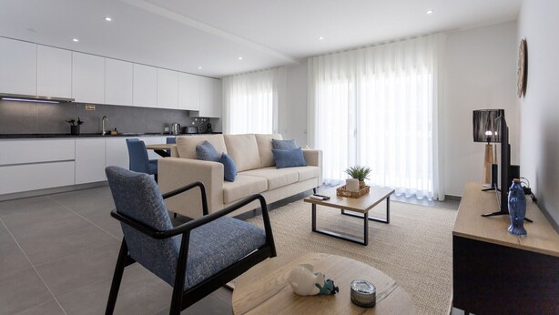 Ein modernes Wohnzimmer mit einer Couch, Sesseln und einem Couchtisch. Es gibt eine Küchenzeile mit Schränken und einer Arbeitsplatte.
