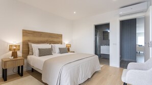 Ein modernes Schlafzimmer mit Holz-Kopfteil, einem großen Bett mit weißer Bettwäsche, zwei Nachttischlampen und Blick ins Bad.