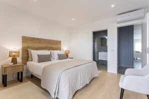 A modern bedroom with a wooden headboard, a large bed with white bedding, two bedside lamps, and a view into the bathroom. - Galeão Sea Apartment - BY SCH (São Martinho do Porto)