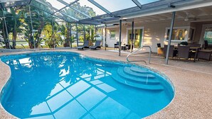 En inomhuspool med väggar av genomskinligt glas, omgiven av solstolar och en möblerad sittgrupp under tak.