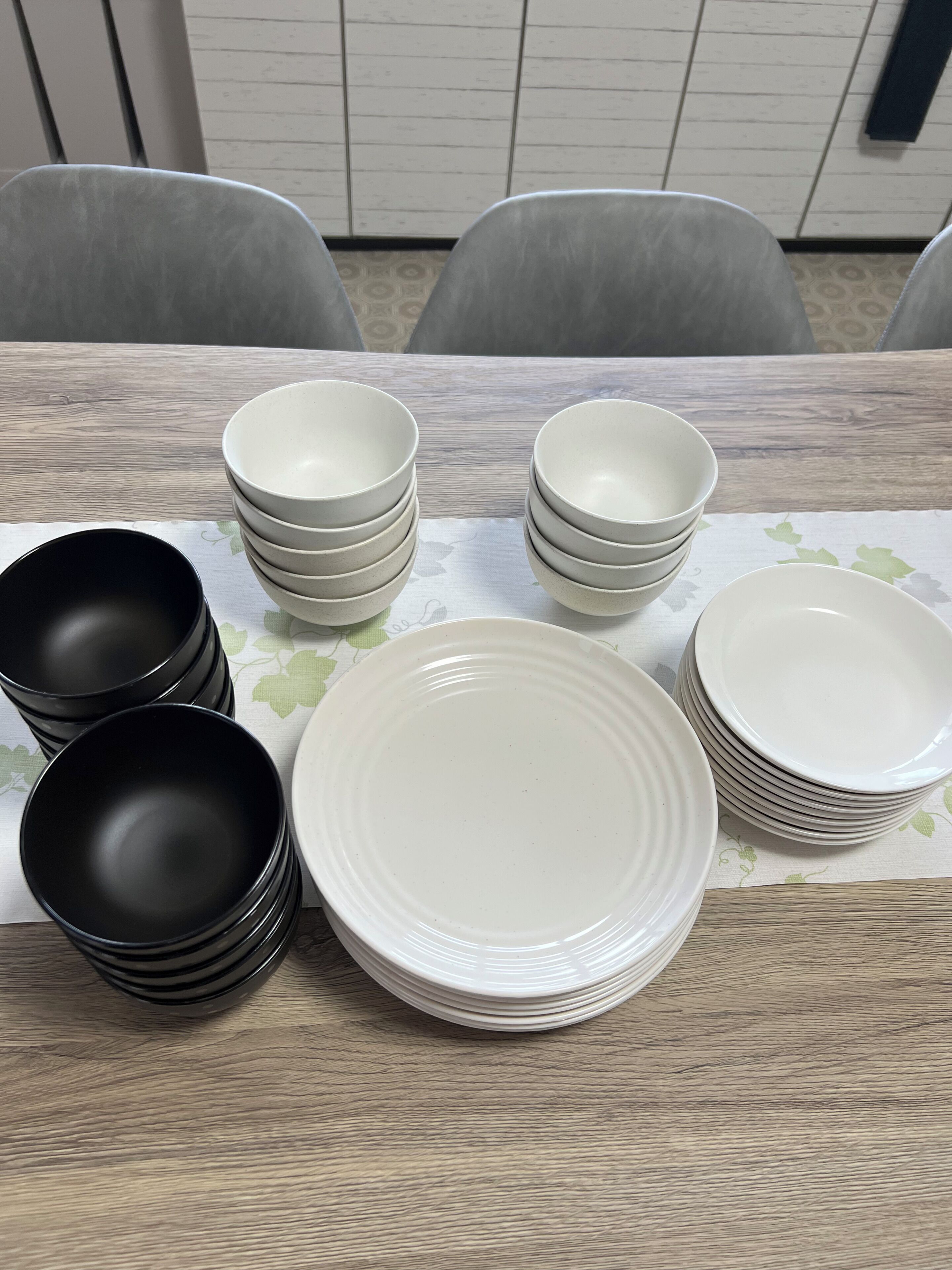 A table with stacks of white and black bowls and plates, a patterned tablecloth, and grey chairs.