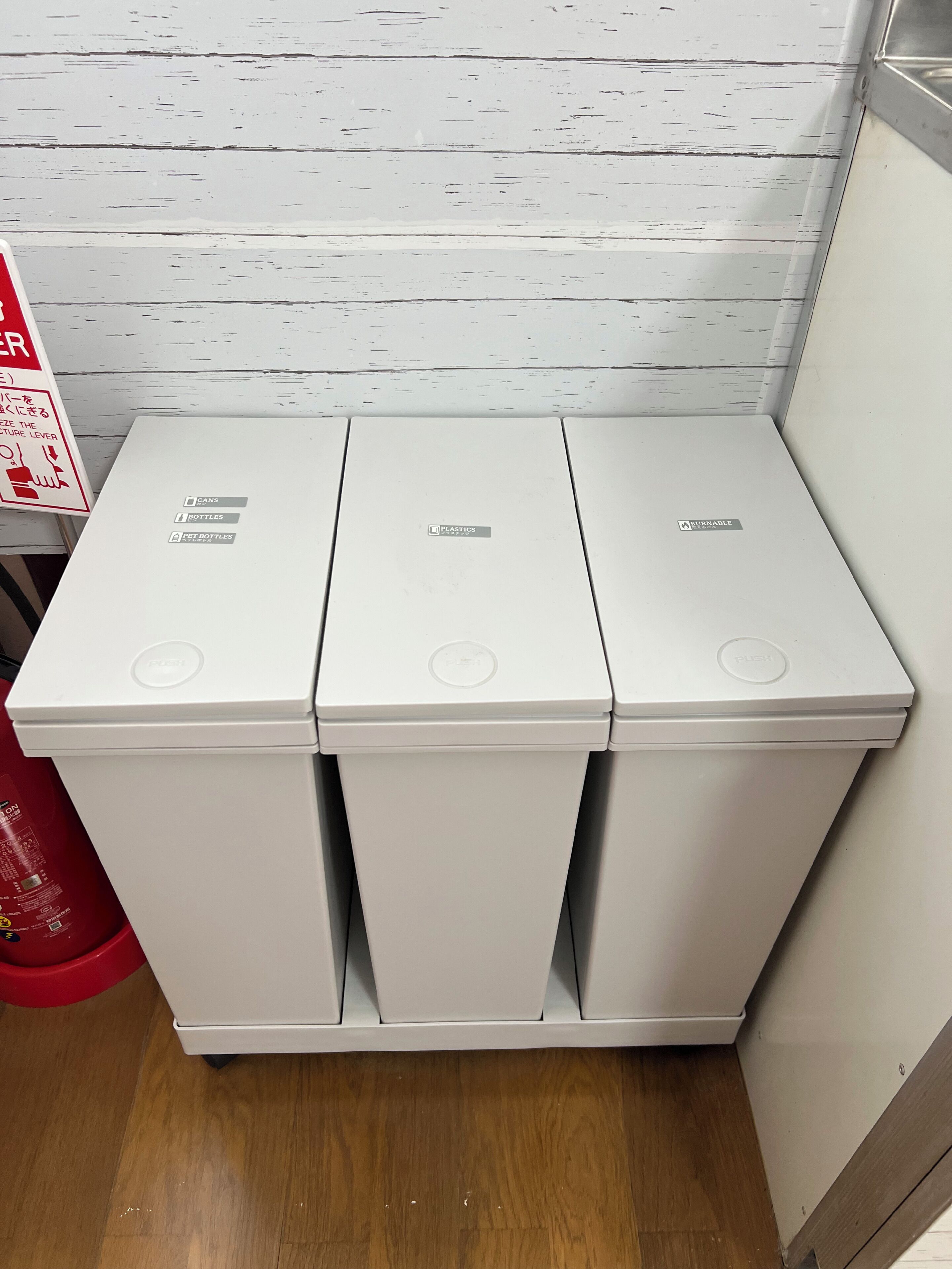 Three white waste bins with recycling symbols, placed against a white wall with a red fire extinguisher to the left.