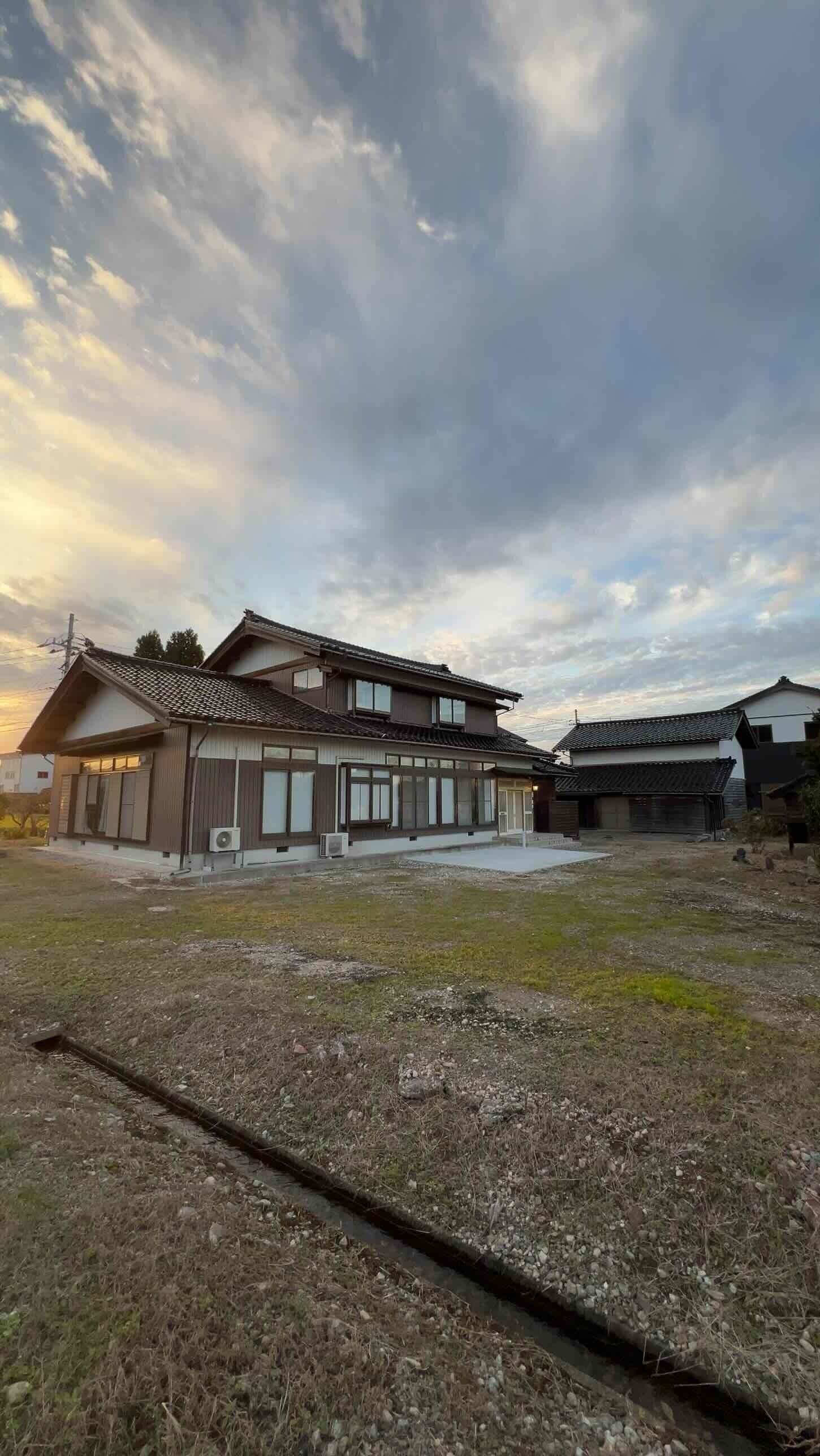 Une maison traditionnelle japonaise, avec un toit recouvert de tuiles, une grande cour ouverte et une zone herbeuse.