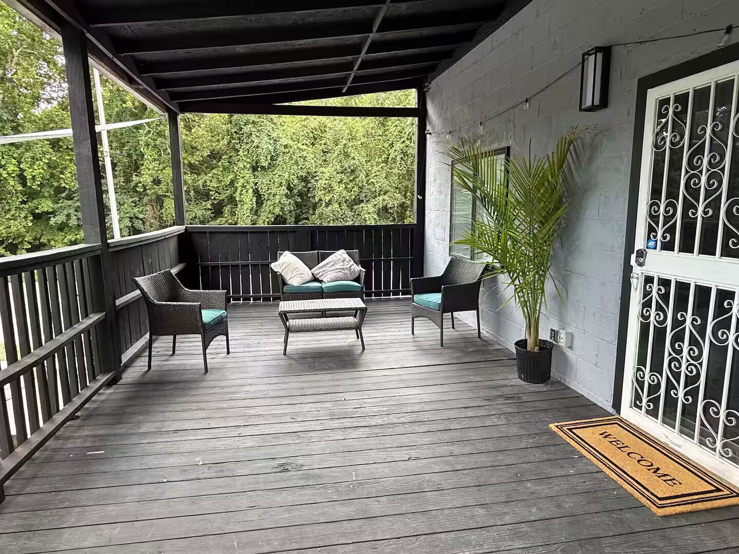 Une véranda couverte avec une terrasse en bois, un canapé avec des coussins, deux fauteuils et une plante en pot.