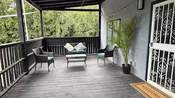 Une véranda couverte avec une terrasse en bois, un canapé avec des coussins, deux fauteuils et une plante en pot.