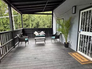 A covered porch with a wooden deck, a sofa with cushions, two chairs, and a potted plant. - KC Group Getaway - Spacious, Very Private - 14+ Ppl - Perfect for Chiefs games! (Independence)