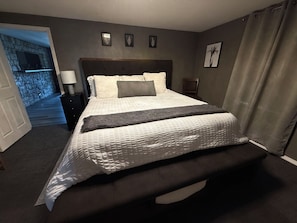 A hotel room with a large bed, a nightstand, a lamp, a chair, and a view of a stone wall and a door leading to another room.