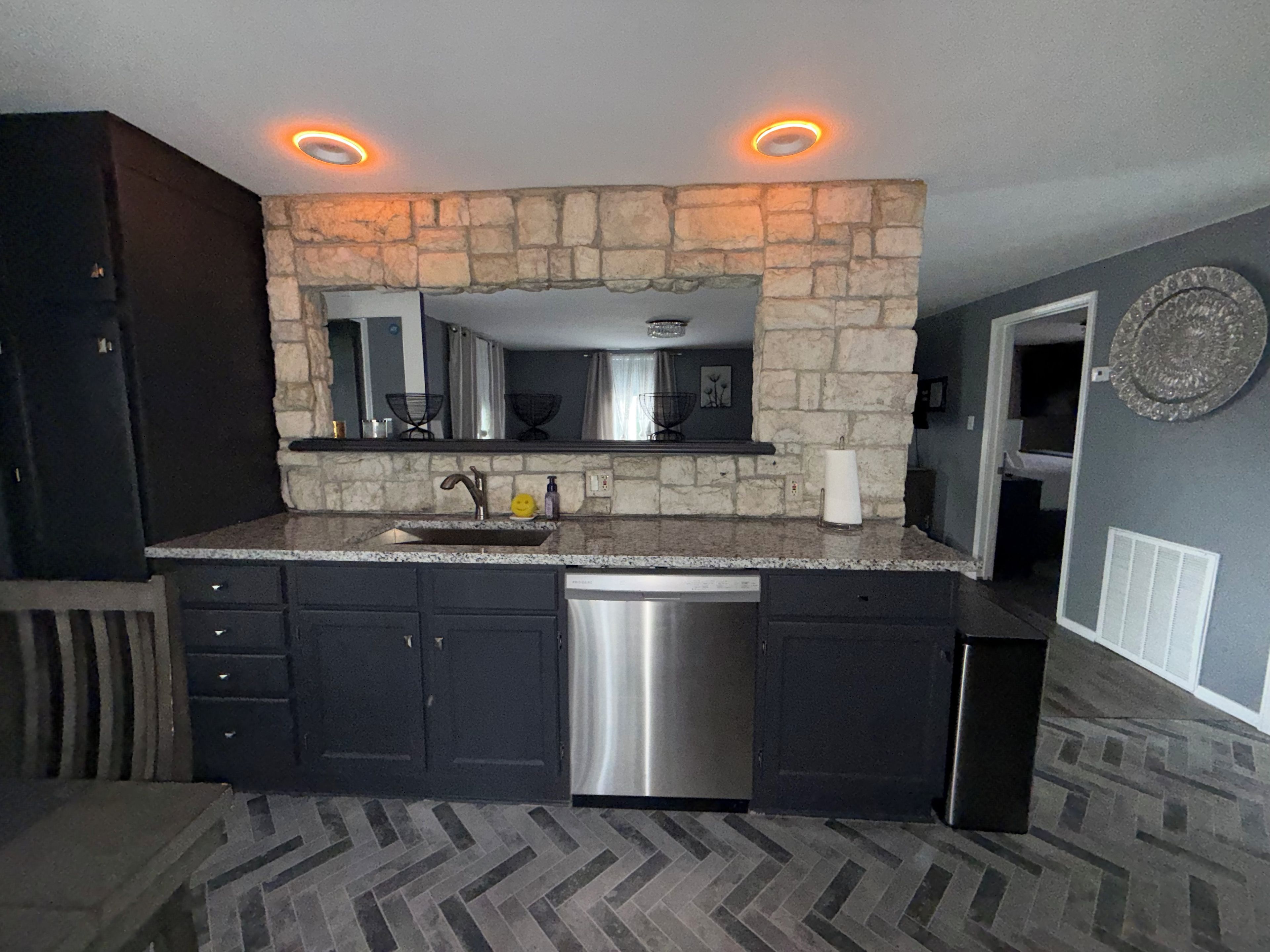 Cozinha com parede de destaque em pedra, armários escuros, lava-louças de aço inoxidável e piso em padrão herringbone.