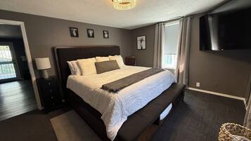 Quarto com cama, mesas de cabeceira, abajur, um banco, televisão e janela com cortinas.