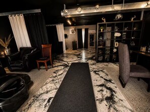A room with a black leather sofa, a wooden chair, a black rug, and a shelf with decorative items. - Modern Retreat / Office / Deck / Massage Chair (Independence)