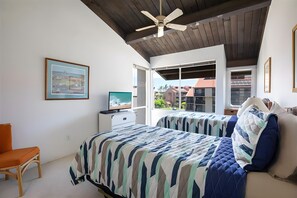 A bedroom with a bed, a desk, a chair, a ceiling fan, and a window with a view of buildings. - Kihei, Hale Kamaole, Spacious 4 BR, Sleeps 12 (Kihei)