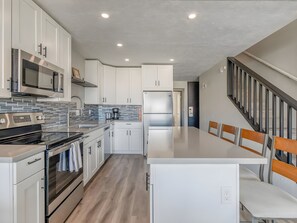 A modern kitchen with stainless steel appliances, white cabinetry, and a wooden dining area. - Kihei, Hale Kamaole, Spacious 4 BR, Sleeps 12 (Kihei)