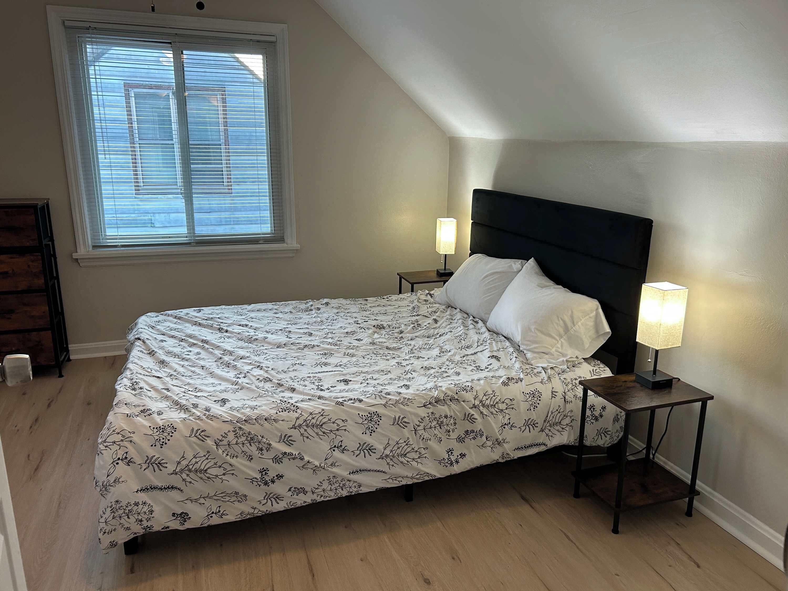 Ein Schlafzimmer mit einem Bett, zwei Nachttischlampen, einem Fenster mit Jalousien und einem Holzboden.