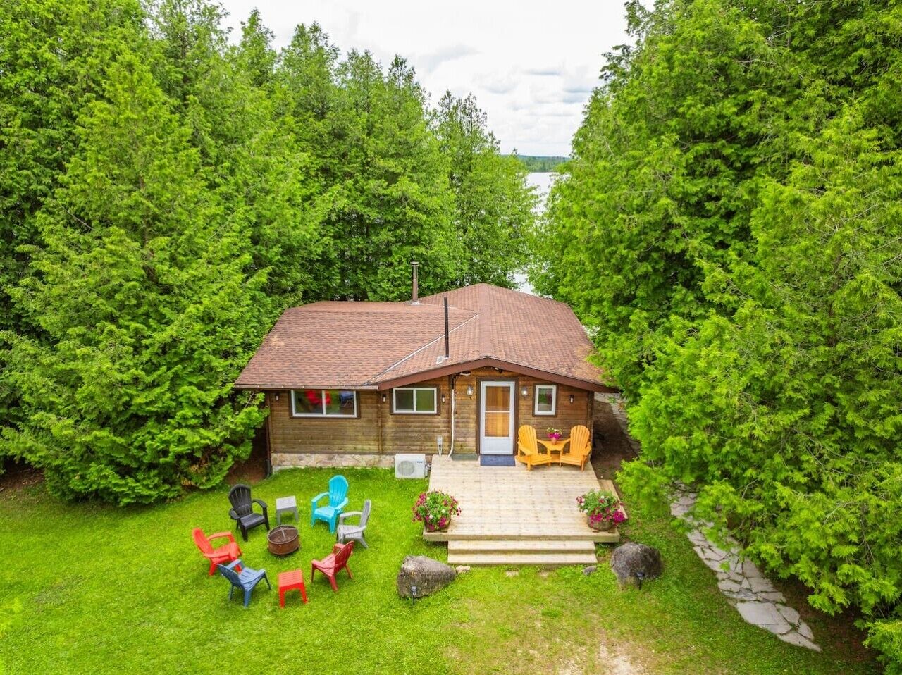 A wooden cabin with a tiled roof, surrounded by lush greenery and a clear view of a lake.