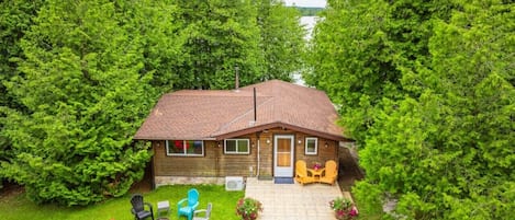 A wooden cabin with a tiled roof, surrounded by lush greenery and a clear view of a lake.