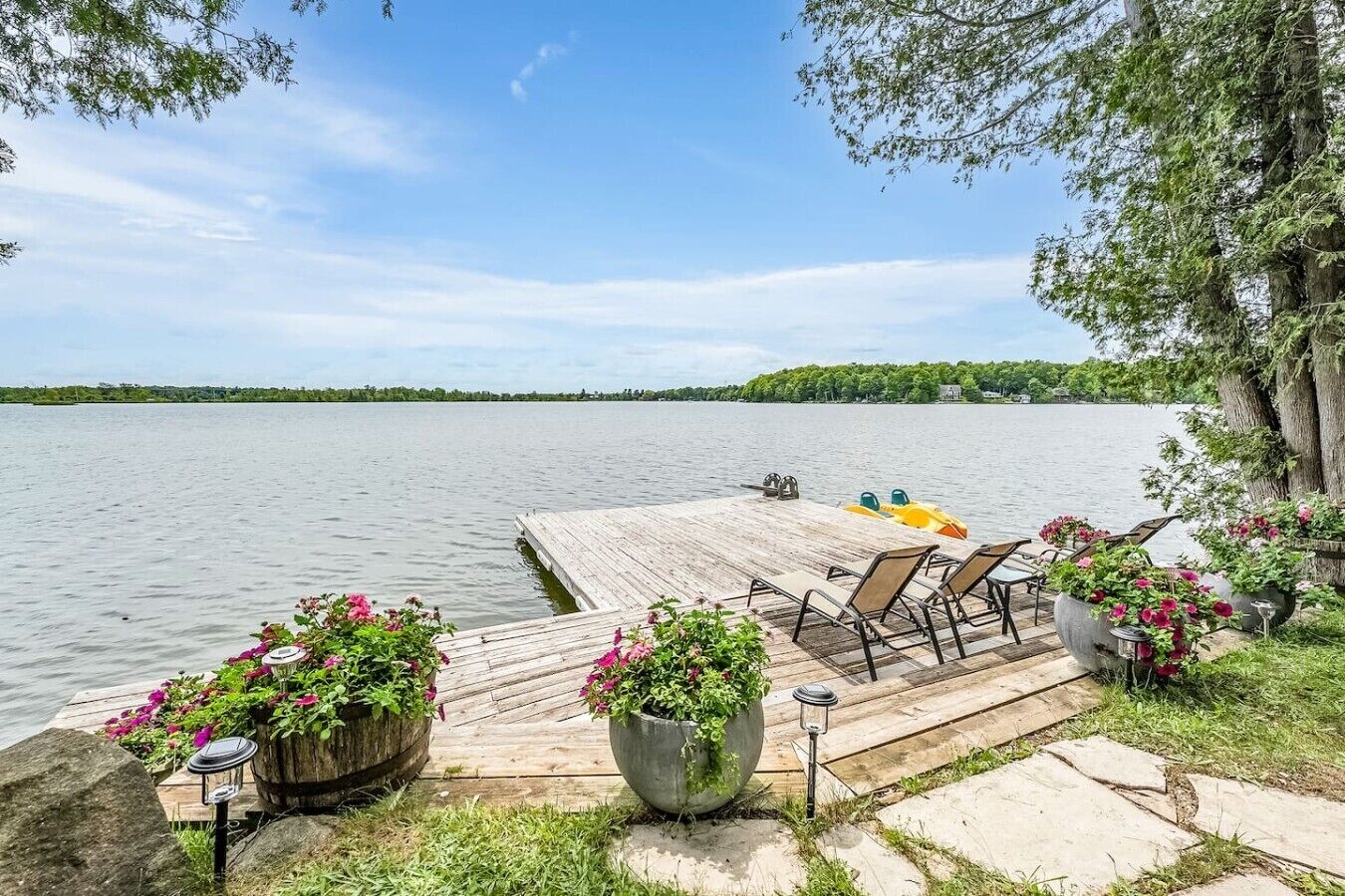 A wooden dock extending into a calm lake with chairs and potted plants, surrounded by trees and a clear sky.