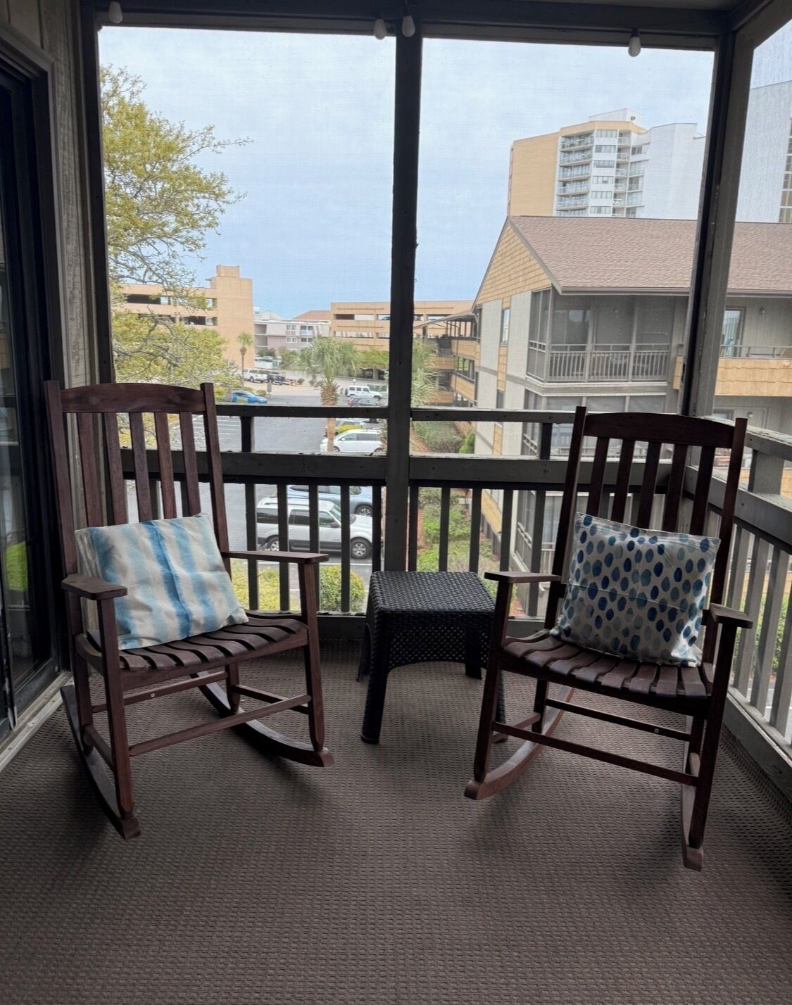 Een balkon met twee houten schommelstoelen en een kleine tafel, met uitzicht op een parkeerplaats en gebouwen.