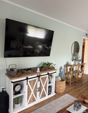 A living room with a mounted TV, a wooden console table with decorative items, a small clock, and a plant. - Marnier’s Cove Condo unit 301 A Myrtle Beach (Myrtle Beach)