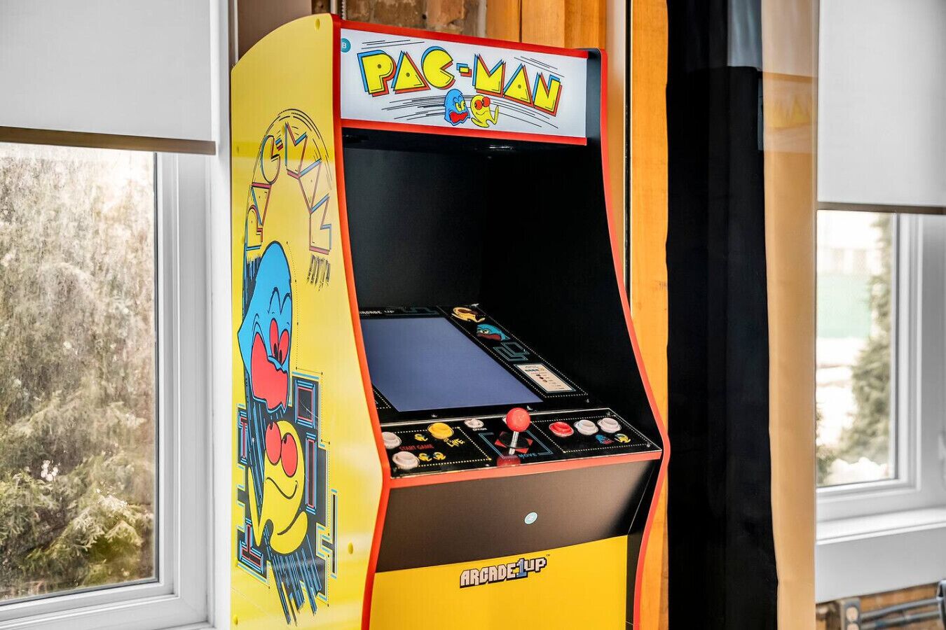 Une borne d’arcade Pac-Man aux couleurs jaune et rouge, placée à l’intérieur, près d’une fenêtre.