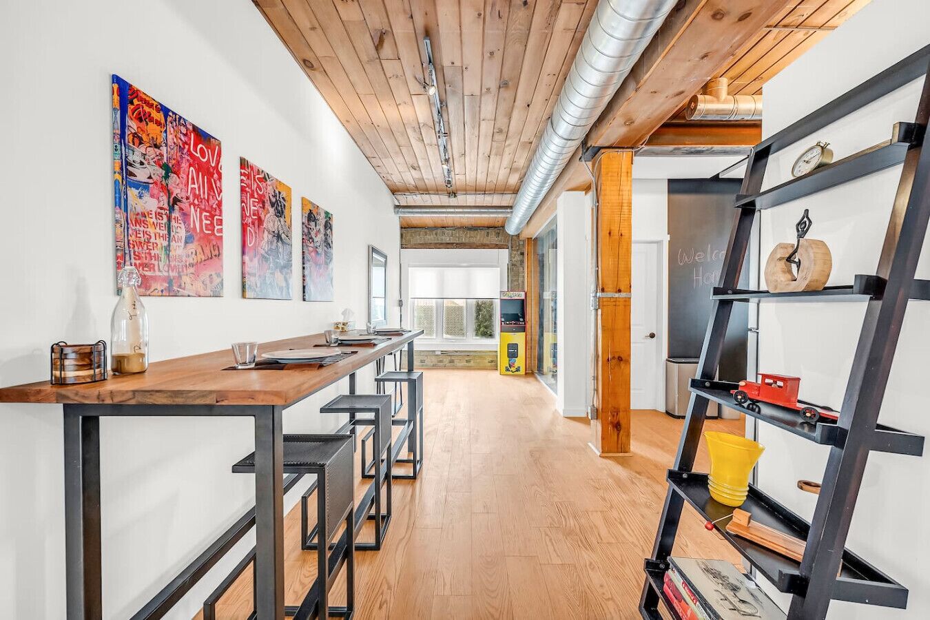 A modern interior with a wooden ceiling, a long table with bar stools, and a black metal shelf with various items.