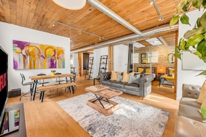 A spacious living room with a wooden ceiling, a large abstract painting, a sofa, a coffee table, and a dining area with chairs and a table. - *Golden Loft*- RARE Downtown 2BR + Parking & Games (Toronto)