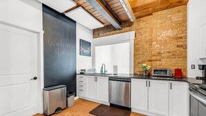A modern kitchen with white cabinets, stainless steel appliances, a chalkboard with 'Welcome Home!' written on it, and a brick wall.