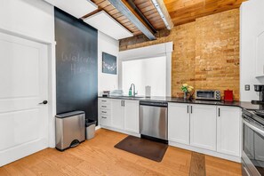 Una cocina moderna con armarios blancos, electrodomésticos de acero inoxidable, una pizarra con la frase '¡Bienvenido a casa!' escrita y una pared de ladrillo.