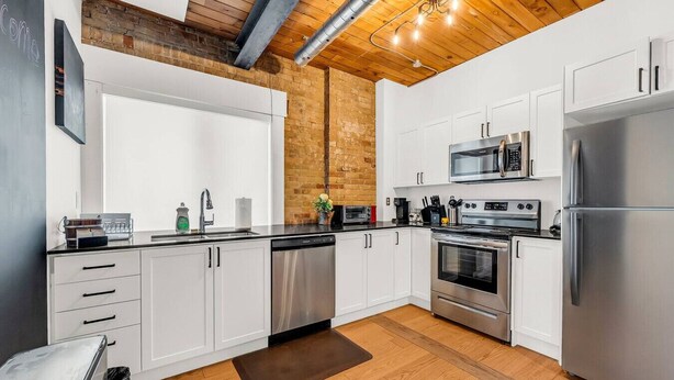 A modern kitchen with white cabinets, stainless steel appliances, a brick accent wall, and a wooden ceiling.