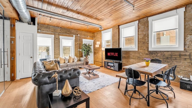 A modern living room with a sofa, dining table, and chairs, a television, and a brick wall.