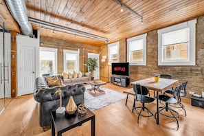 A modern living room with a sofa, dining table, and chairs, a television, and a brick wall. - *Golden Loft*- RARE Downtown 2BR + Parking & Games (Toronto)