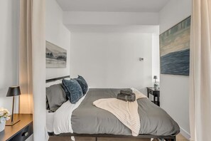 Una camera da letto con un letto, cuscini, una coperta, un comodino con una lampada e un quadro appeso al muro.