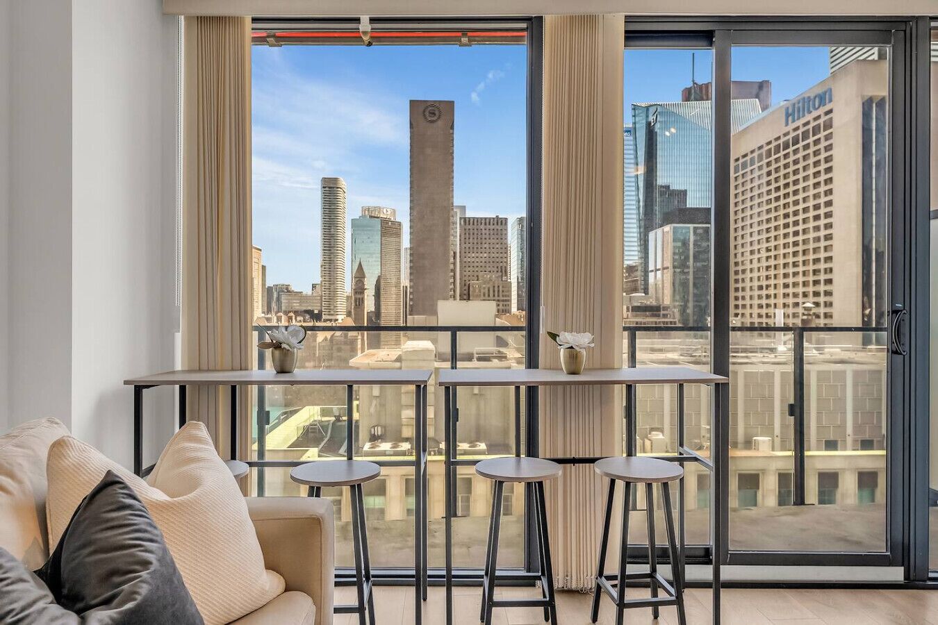 A modern living room with a city view, a sofa, a coffee table, and two bar stools.