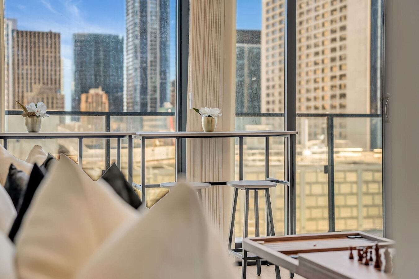 A modern hotel room with a large window, a view of tall buildings, a small table with a chess set, and a stool.