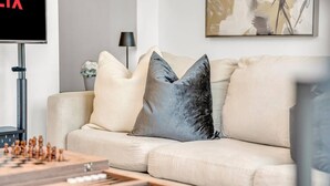 Ett vardagsrum med en beige soffa, två kuddar, en tv och ett schackbräde.