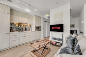 Un espacio de estar moderno con cocina, un televisor de pantalla plana montado en un mueble, una mesa de ajedrez y un sofá con cojines.